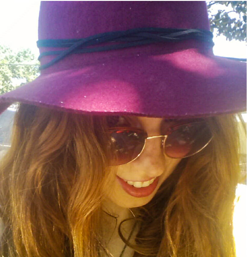 Photo of the artist: peering over brown-tinted sunglasses, maroon hat and red hair obscuring most of her face. A crooked smile means she might be up to something.